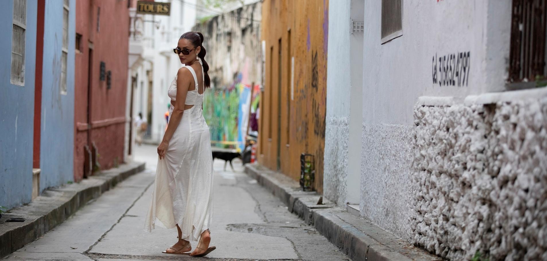 Shay Mitchell looking back over her shoulder while wearing a white matching set surrounded by colorful buildings in South America.